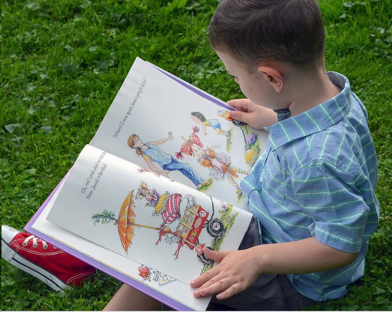 A boy is reading a book outside on the grass.